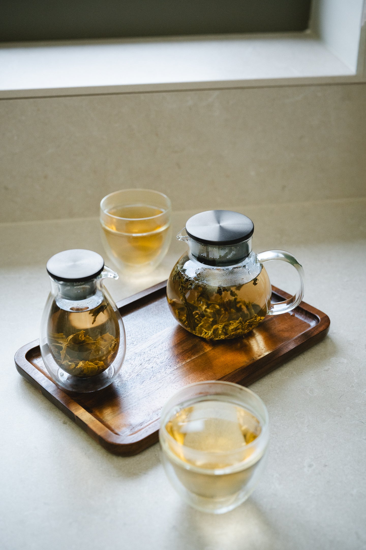 Tea-making setup with two different styles of glass teapots filled with brewed loose tea, two glass cups, and a wooden tray on a light surface.