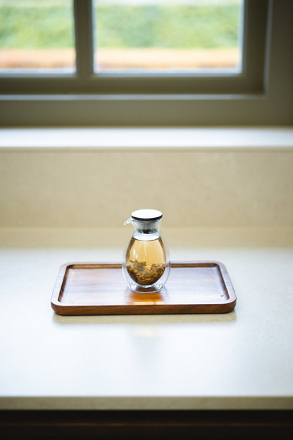 Small glass handle-less teapot filled with brewing tea on a wooden tray in front of a window.