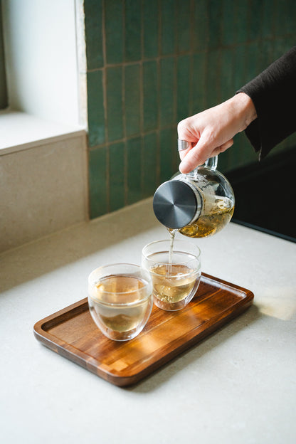 Person pouring tea from a glass teapot with a filter in the lid into two glass cups on a wooden tray.