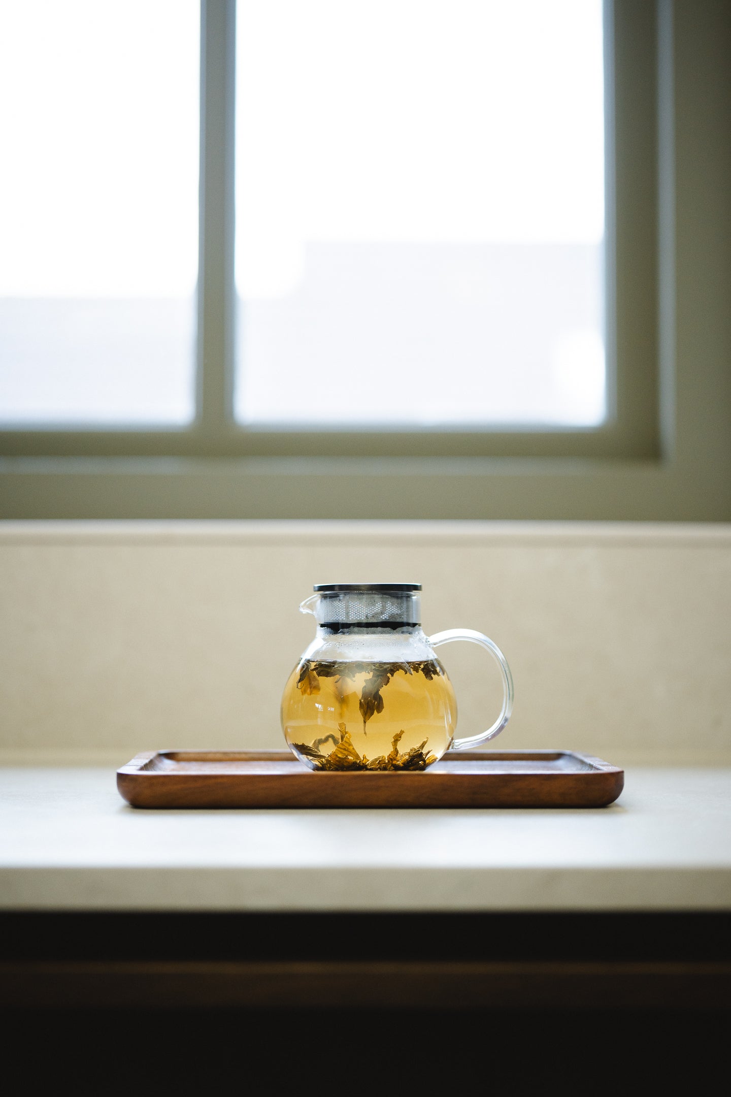 Clear glass tea pot with a metal lid filled with brewing tea on a wooden tray in front of a window