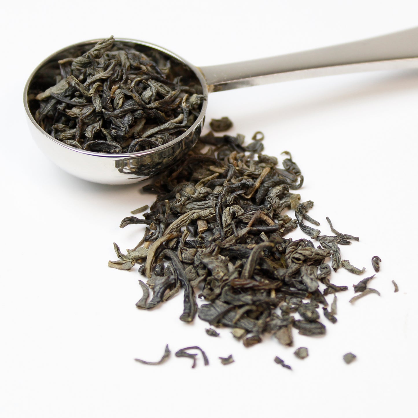 Loose leaf Chun Mee green tea leaves spilling out of a stainless steel teaspoon on a white background