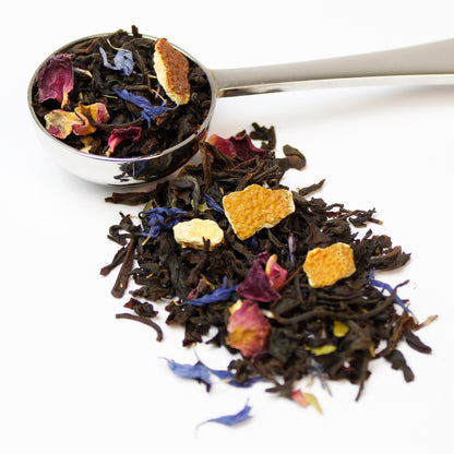 Black tea leaves with orange pieces, lime leaves and flower petals with a stainless steel spoon on a white background.