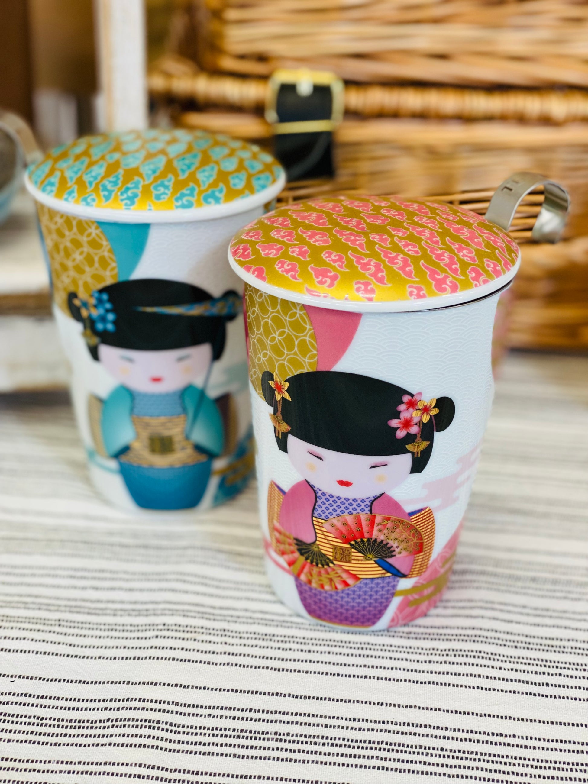 Two handle-less tea mugs with colorful designs of Japanese geisha characters on a striped surface.