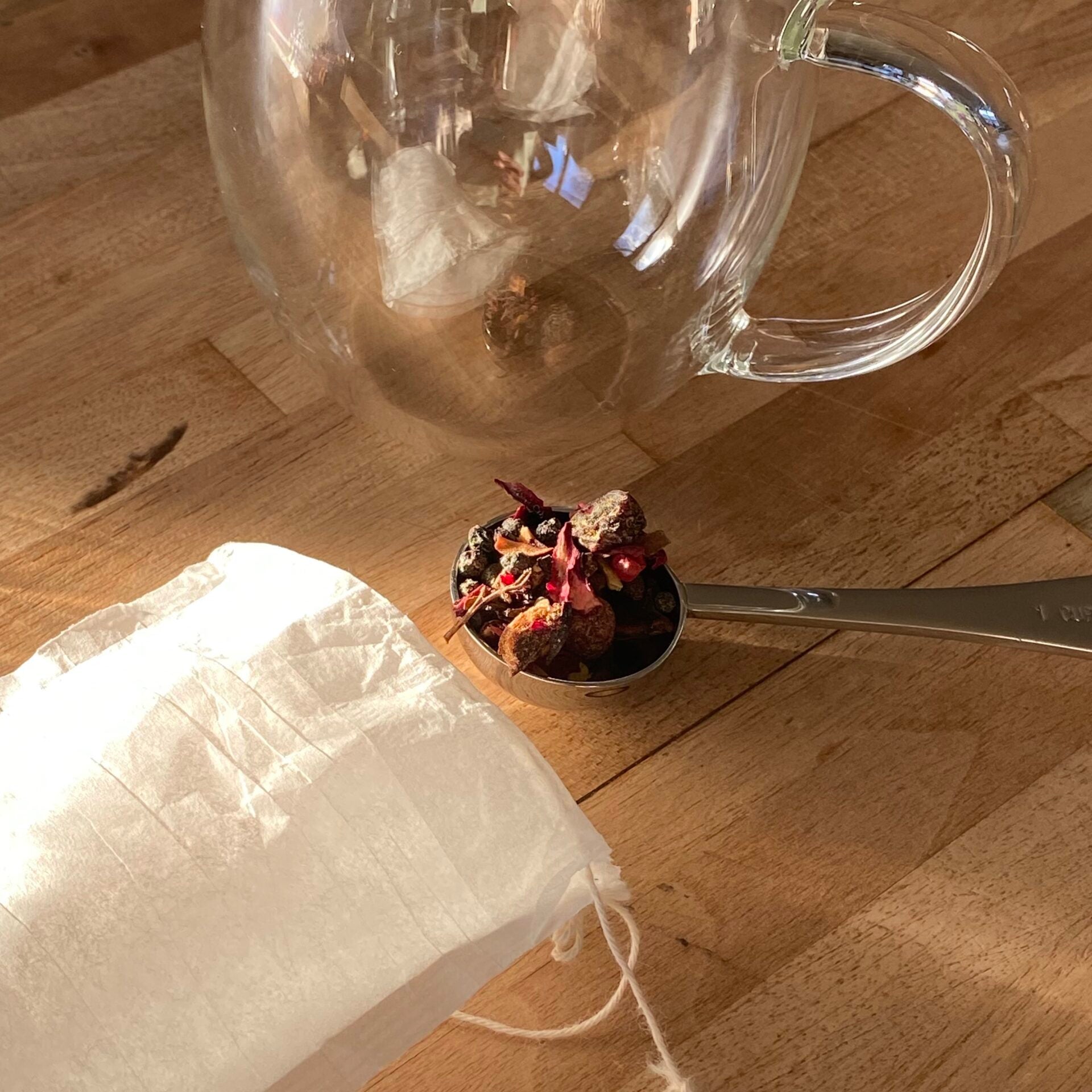 Clear glass cup next to a spoonful of herbal tea and a compostable paper tea filter on a wooden surface