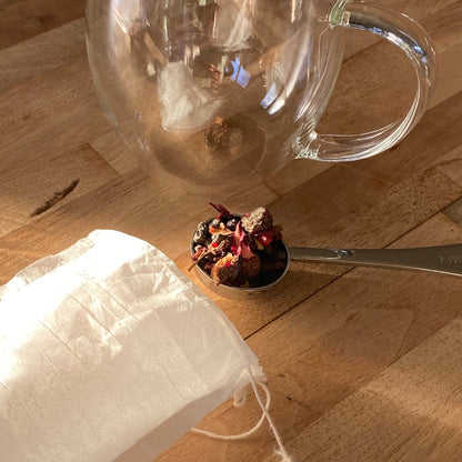 Clear glass cup next to a spoonful of herbal tea and a compostable paper tea filter on a wooden surface
