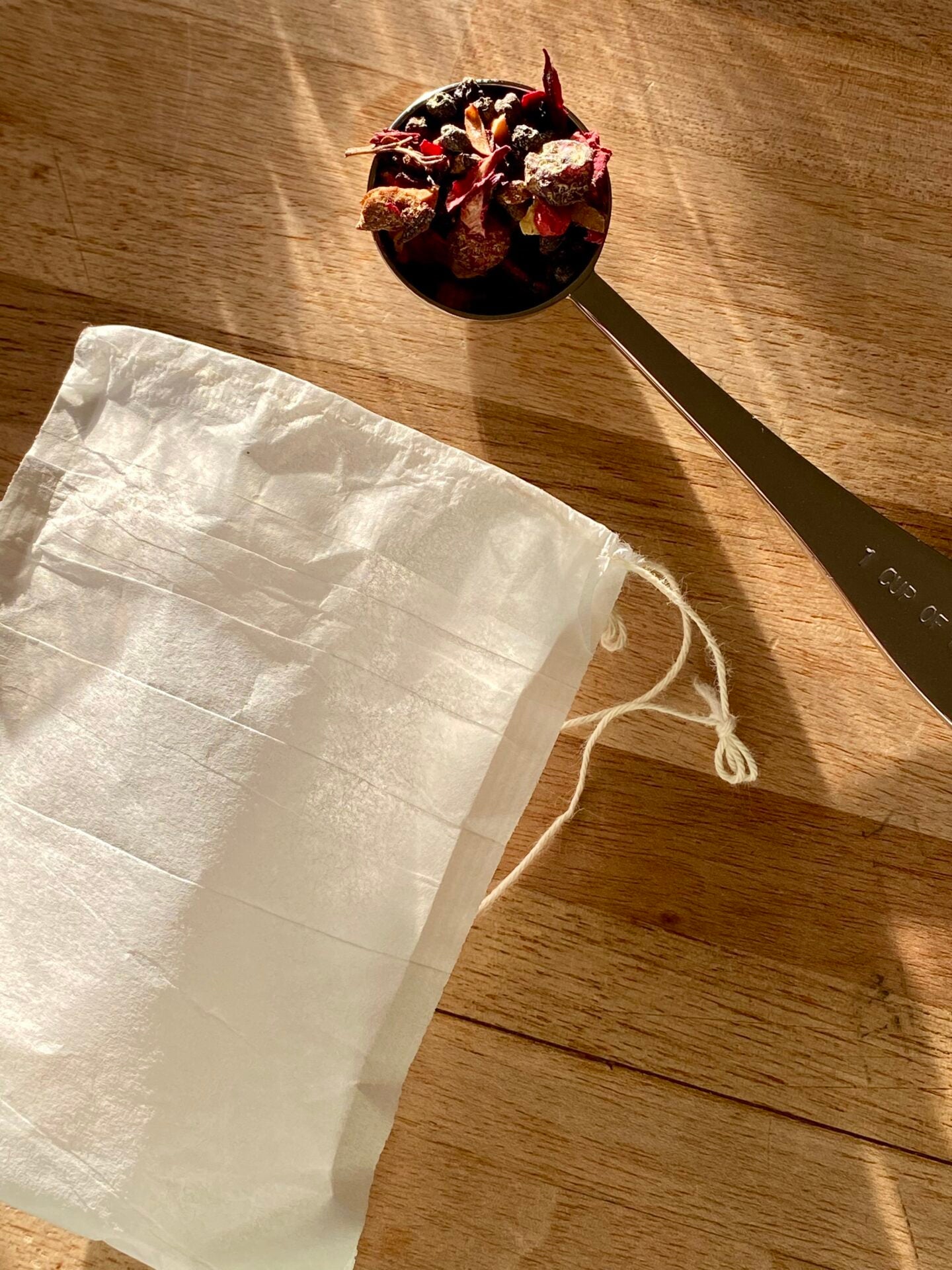 White compostable paper tea filter with drawstring on a wooden surface next to a stainless steel spoon of herbal tea.