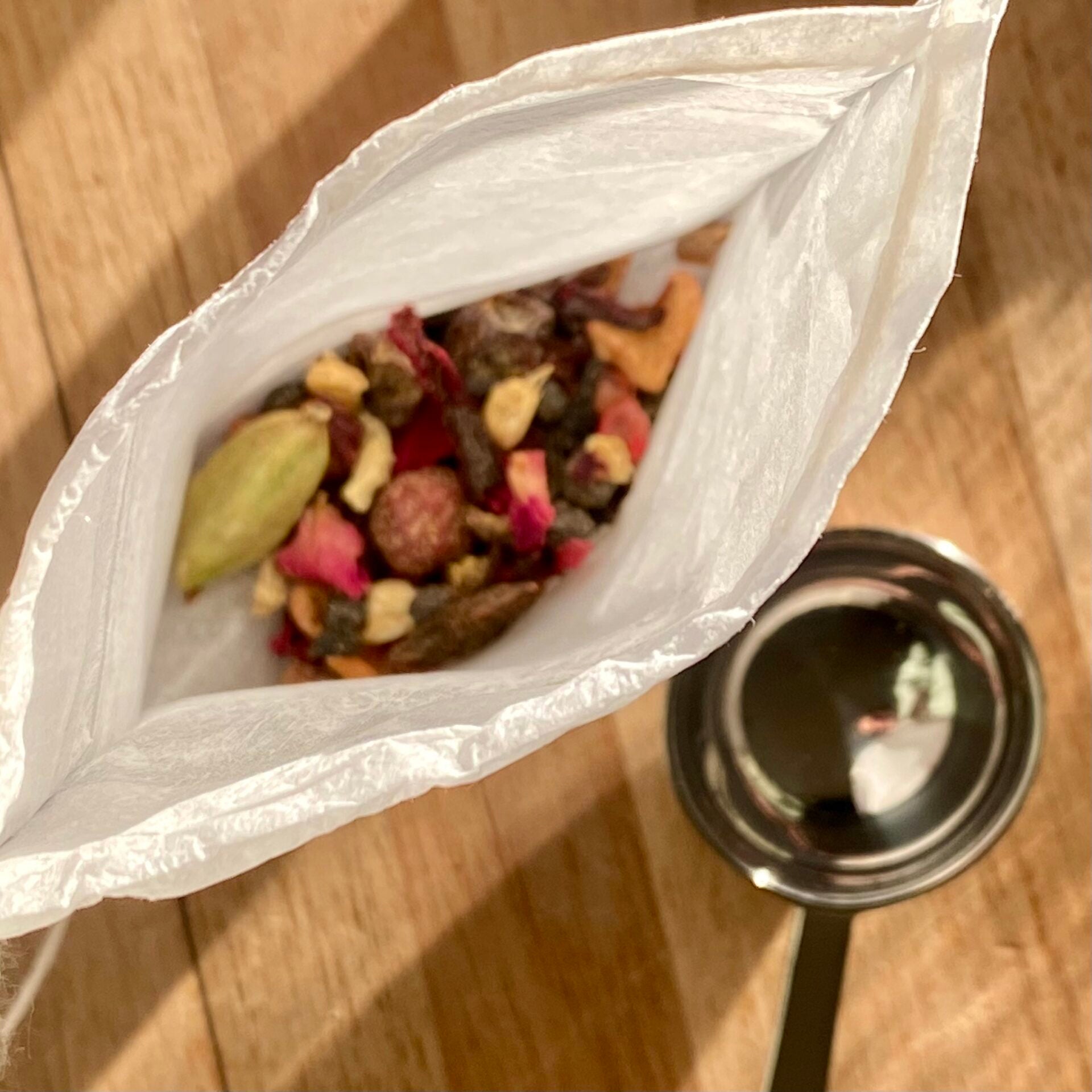 White compostable paper tea filter with drawstring filled with herbal tea on a wooden surface next to a stainless steel spoon.