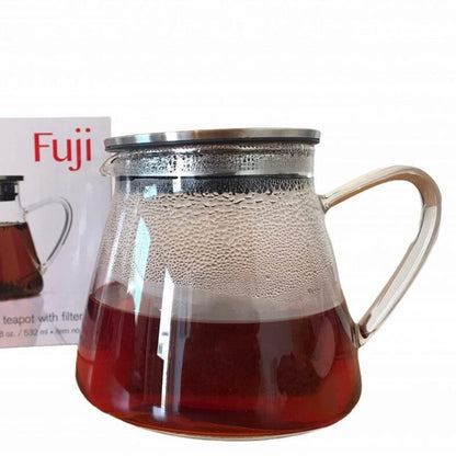 Glass teapot with metal lid and handle filled with hot tea next to its branded box on a white background.