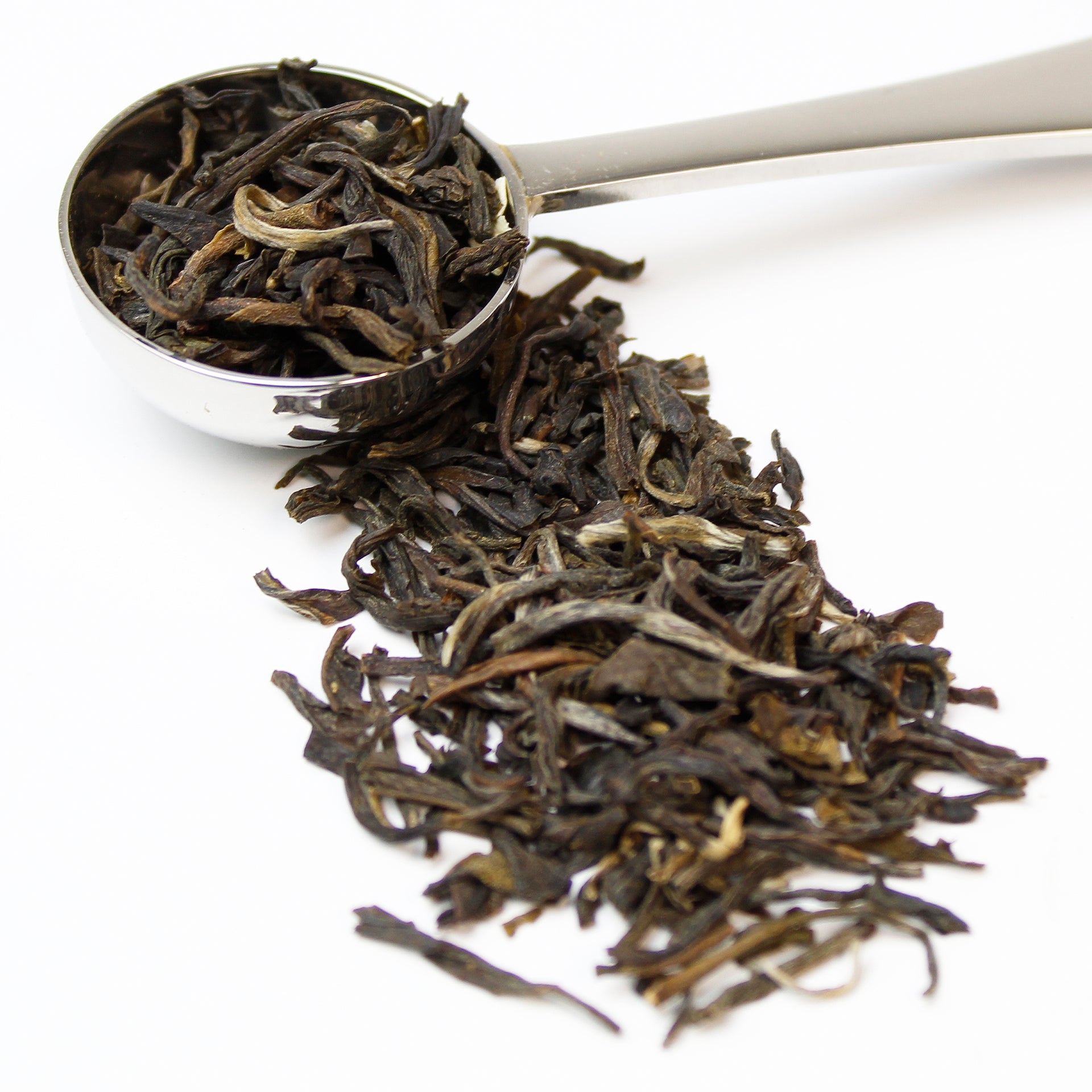 Loose leaf tea leaves in a silver teaspoon on a white background.