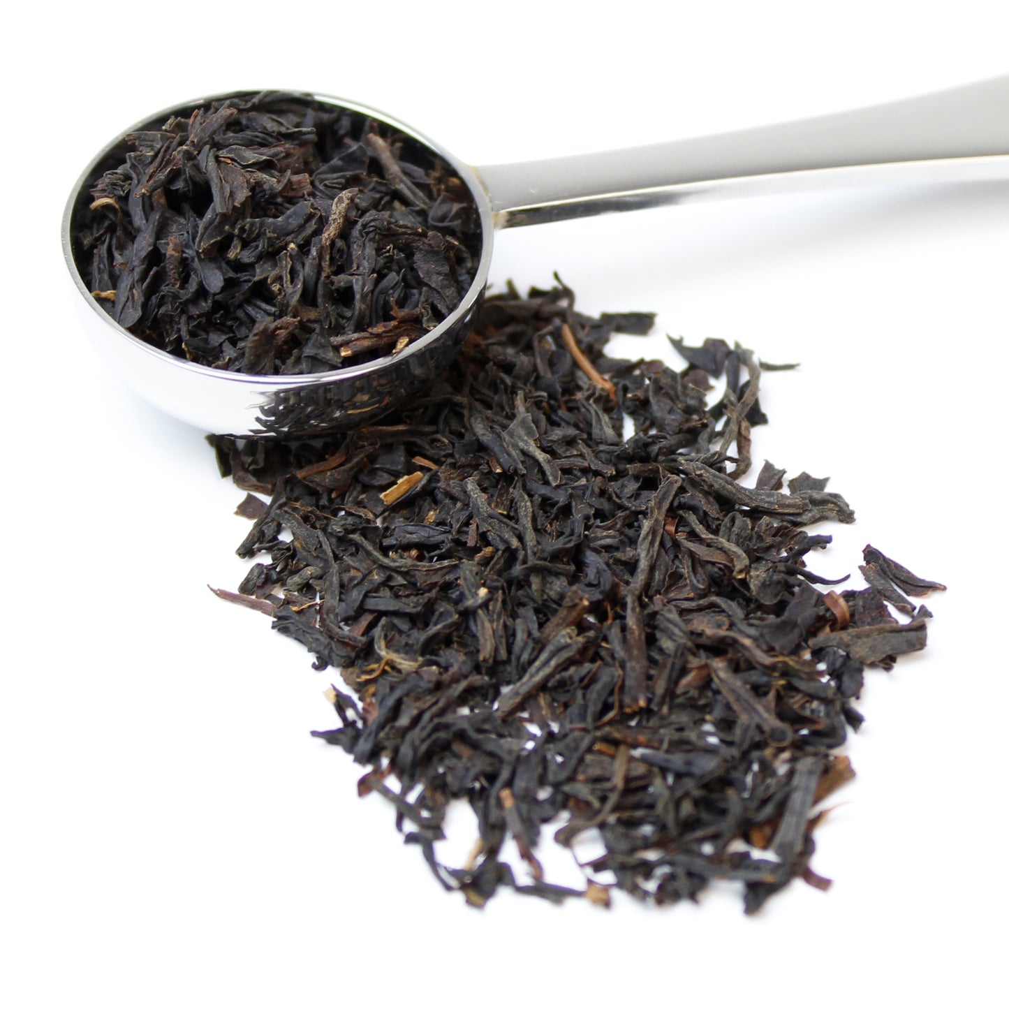 Black loose leaf tea spilling from a silver teaspoon and sitting on a white background.