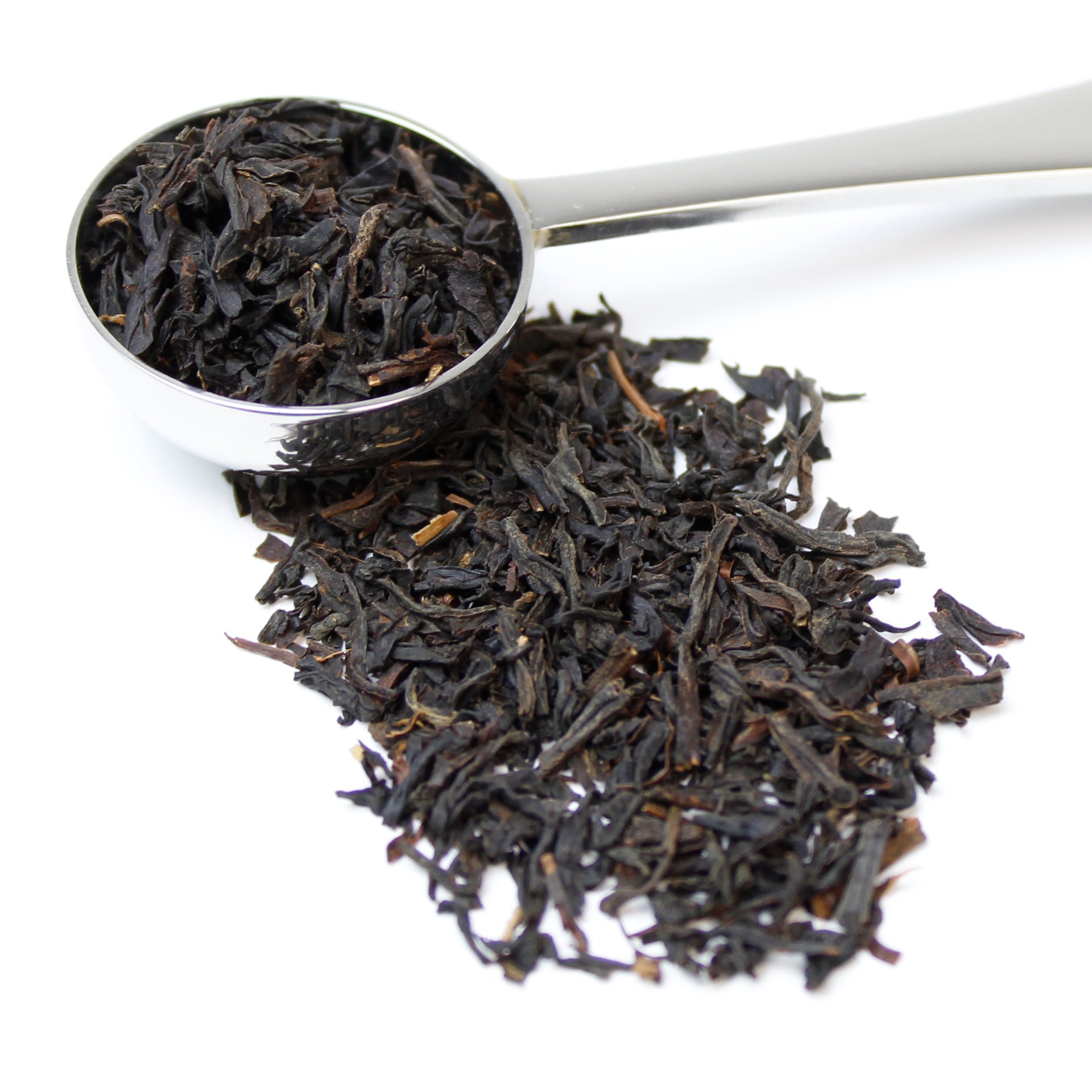 Black loose leaf tea spilling from a silver teaspoon and sitting on a white background.