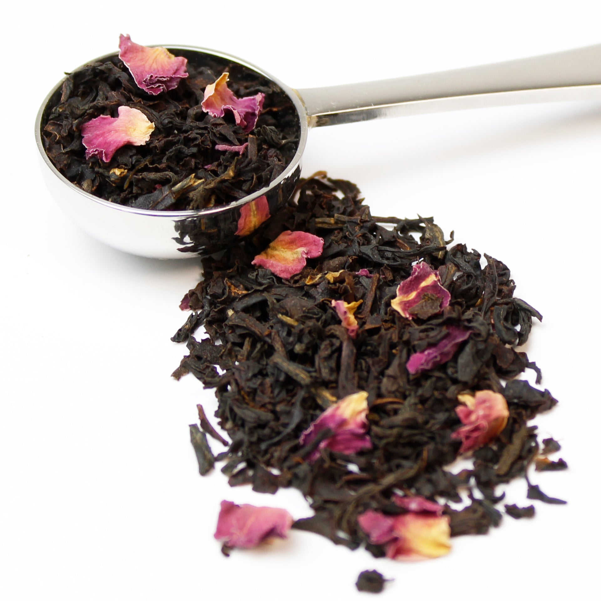A silver teaspoon filled with black tea with rose petals on a white background.