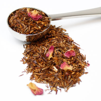 Dried and cut Rooibos leaves with pink rose petals in a stainless steel teaspoon on a white background