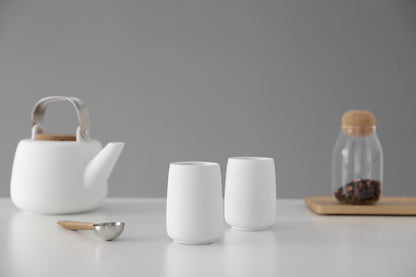 Lifestyle image of white teapot, two porcelain cups, and a jar of tea on a grey background