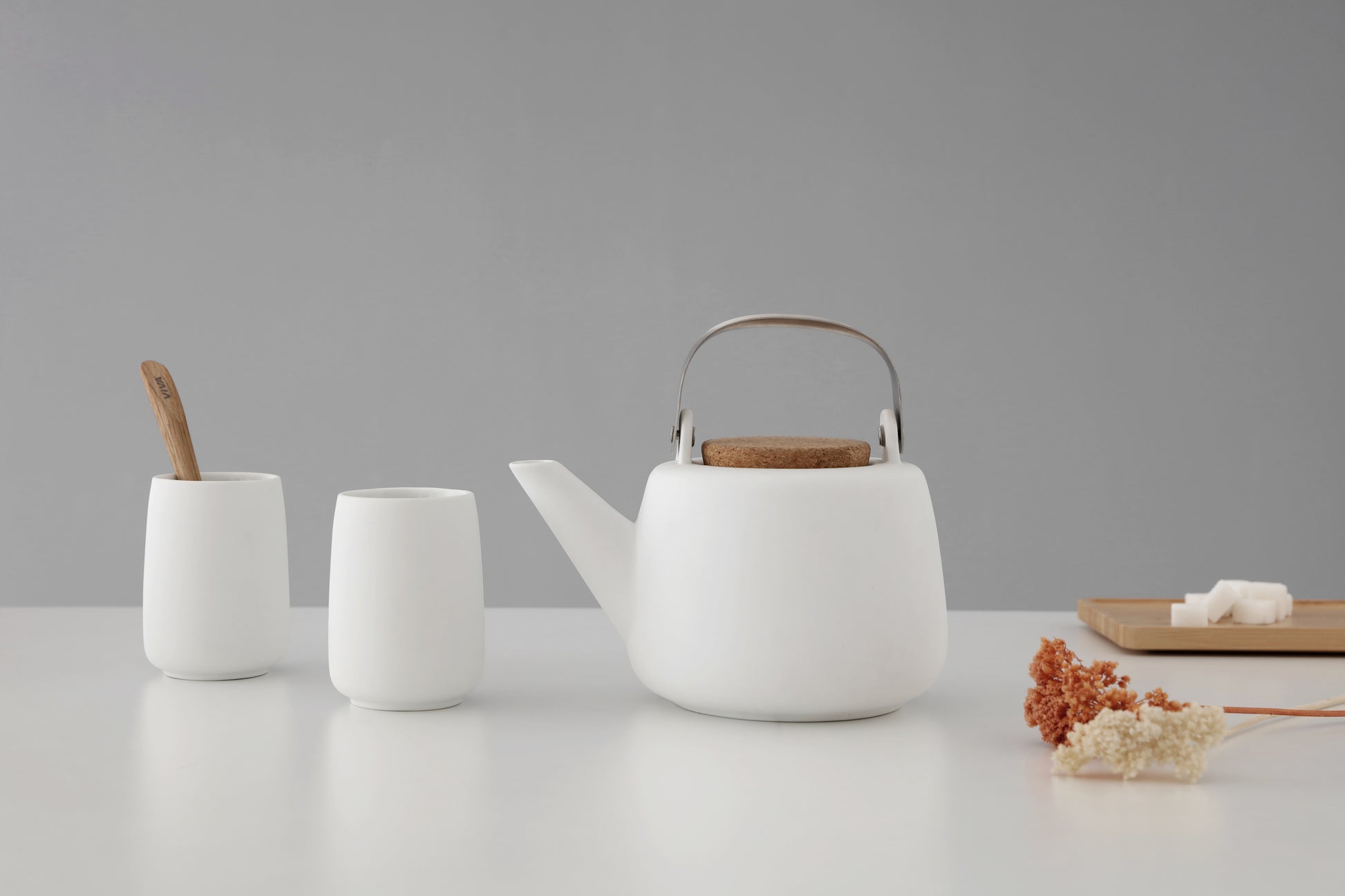 Lifestyle image of white teapot with cork lid, two white porcelain cups, and a small wooden tray with decorative items on a grey background.