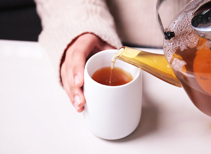 Tea being poured from a glass teapot into a porcelain white cup on a light surface.